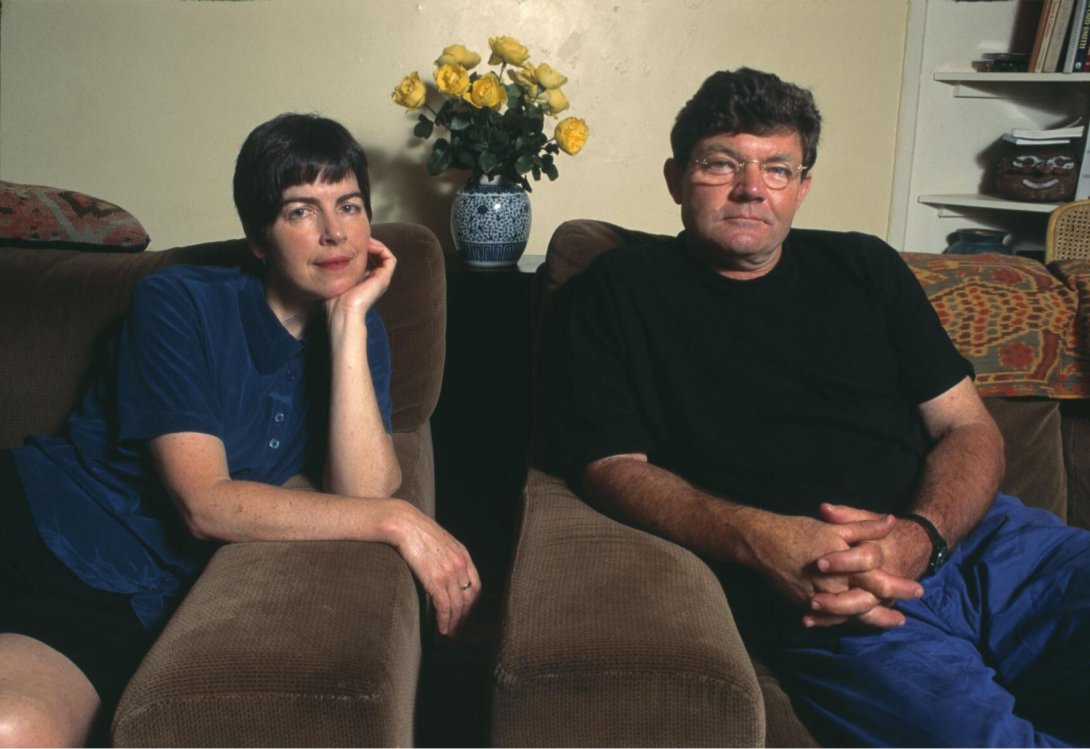 A woman and a man sitting in matching brown armchairs. In the background is  vase with yellow flowers in it, and a white shelf with some books on it.